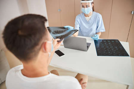 Professional young doctor in protective mask giving an x-ray image to her patient and looking him in...
