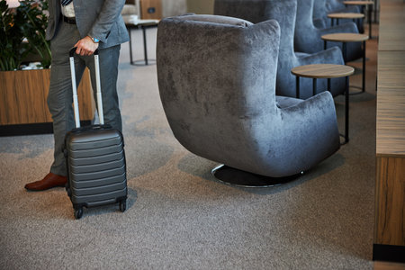 Cropped photo of a person holding a handle of a wheeled suitcase while standing near a padded seatの写真素材