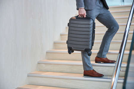 Cropped photo of a person wearing grey pants and brown shoes standing with a suitcase