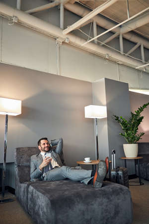 Glad businessperson with a phone lying on his back on a couch between two lamps and smiling with satisfactionの写真素材