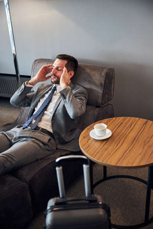 Waist-up photo of a worn-out entrepreneur rubbing his temples next to a coffee cup and carry-onの写真素材