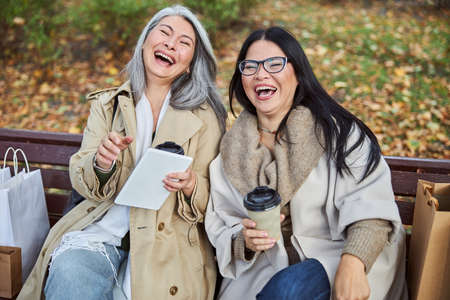 Joyful female friends holding takeaway coffee cups and laughing while using tablet computer outdoors