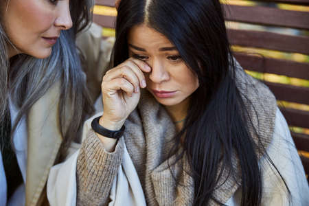 Upset lady sitting on bench and wiping tears while friend looking at her with sympathyの写真素材