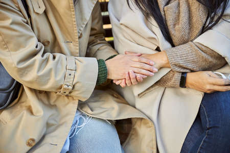 Close up of woman in trench coat holding hand of female friend while sitting on benchの写真素材