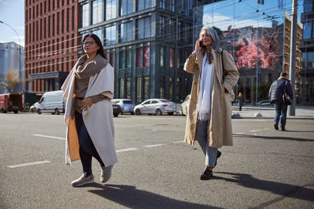 Stylish lady talking on cellphone and smiling while crossing the road with charming brunette womanの写真素材