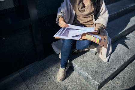 Close up of businesswoman holding folder with papers while sitting on steps outdoorsの写真素材