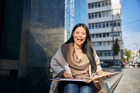 Beautiful lady holding smartphone and laughing while working with documents on the streetの写真素材