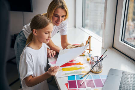 Attractive smiling pleased blonde young Caucasian woman looking at a drawing in her daughter hand