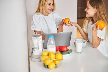 Smiling pleased female parent looking at her beautiful generous daughter treating her to the citrus fruitの写真素材
