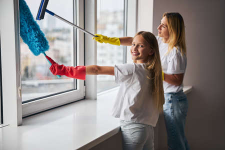Smiling cute girl and her mother using a squeegee and a microfiber duster for window cleaningの写真素材