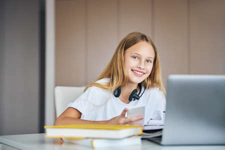 Glad pupil having a smile on her face while grabbing a phone from a desk and working on school exercisesの写真素材