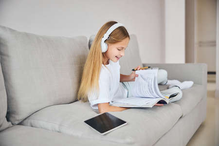 Adolescent person wearing wireless headphones and listening to some songs from the tablet while turning book pagesの写真素材