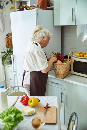 Nice senior lady in apron standing by the kitchen counter with fresh vegetables, berries and fruitsの写真素材