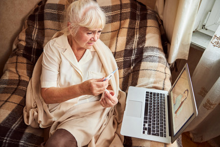 Upset elderly lady sitting in armchair and holding digital thermometer while talking with female doctor through video callの写真素材