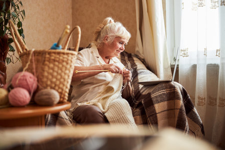 Nice elderly woman watching online knitting lesson on notebook while sitting in armchair and knitting scarf at homeの写真素材