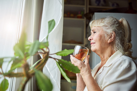 Nice elderly lady looking out the window and smiling while holding cup of hot drinkの写真素材