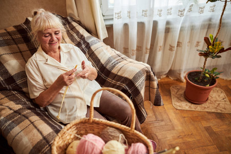 Nice senior lady looking at needles with yarns and smiling while resting in comfortable chair at...