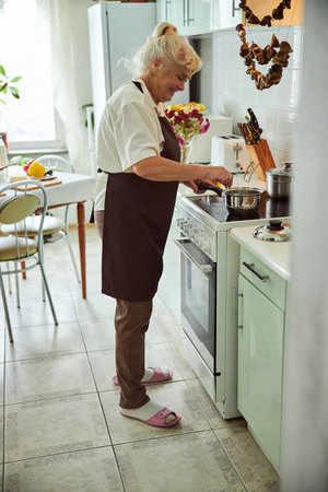 Smiling elderly lady in apron heating up food on the stove in kitchen