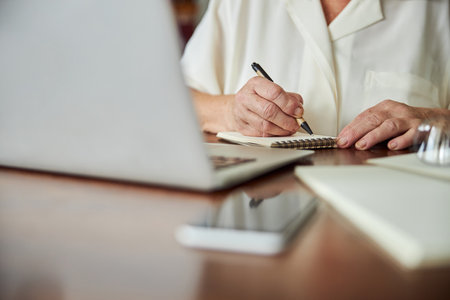 Close up elderly lady making notes while working on modern notebook at home