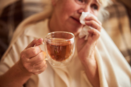 Close up of sick old lady holding cup of hot herbal drink and using handkerchiefの写真素材
