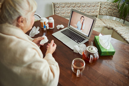 Elderly lady having online consultation with doctor while sitting at the table with laptop and medicamentsの写真素材