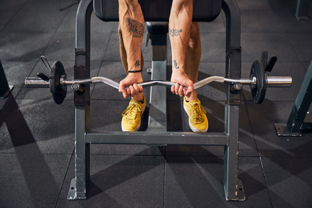 Cropped photo of a strong tattooed man performing a barbell curl on the weight benchの写真素材
