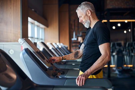 Side view of a handsome focused serious bearded gray-haired male athlete turning on the treadmill