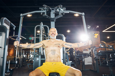 Handsome cheerful Caucasian gray-haired male athlete using the seated fly machine in a fitness...