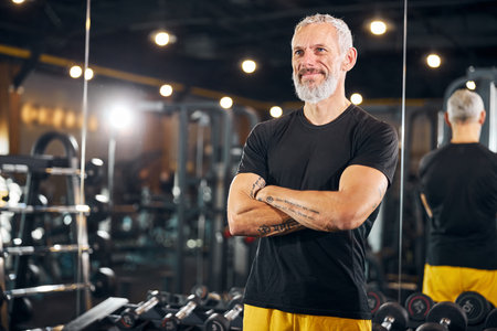 Attractive smiling dreamy muscular gray-haired Caucasian fitness instructor with folded arms standing at the gymの写真素材