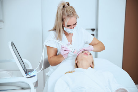 Concentrated beautician in a medical mask doing a cosmetic procedure for improving her patient skin elasticityの写真素材