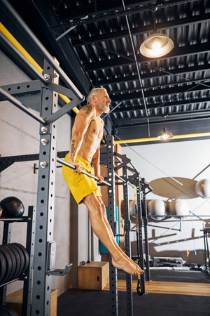 Side view of a barefoot shirtless athletic man in shorts exercising on the pull-up bar