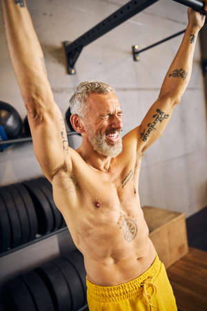 Laughing sportsman hanging with arms fully extended from the horizontal bar during the strength...