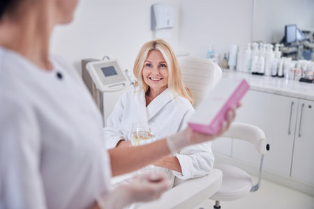 Charming elegant female in white plush bathrobe sitting on the chair and holding glass of water while listening her cosmetologist in beauty centerの写真素材