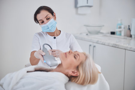 Waist up portrait of beautician applying ultrasound special beautician device for spa treatment and anti aging massage on neck in beauty centerの写真素材
