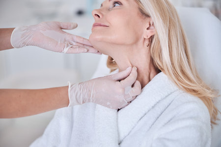 Cropped head portrait of charming blonde Caucasian woman in white plush bathrobe sitting on beauty couch while doctor examining skin on neck in beauty centerの写真素材
