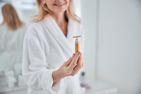 Cropped head portrait of happy smiling woman in white plush bathrobe holding roller massager in hand while standing in front of mirrorの写真素材