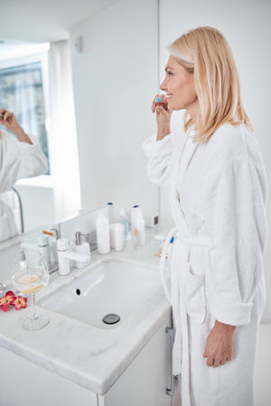Side view portrait of blonde Caucasian beautiful woman in white plush bathrobe looking to the mirror in bathroom while brushing her teethの写真素材
