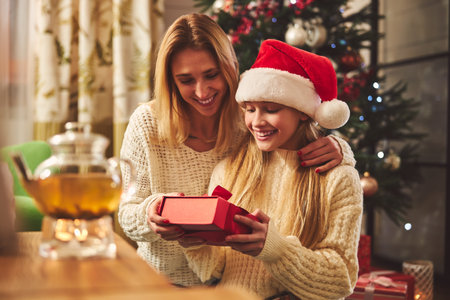 Cheerful attractive woman with female teenager are sitting at home near Christmas tree and opening presentsの写真素材