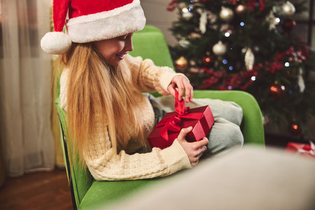 Happy pretty girl is sitting in decorated living room and unwrapping presents during winter holidaysの写真素材
