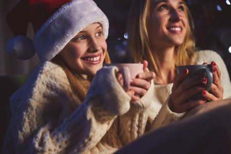 Smiling pretty teenager and parent are drinking hot tea together at home in holy eveningの写真素材