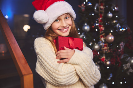Portrait of joyful beautiful girl in Santa hat hugging with love box with gift on New Year Eveの写真素材