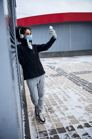 Adult in a medical mask showing her hand in a greeting gesture to the frontal camera of a smartphoneの写真素材