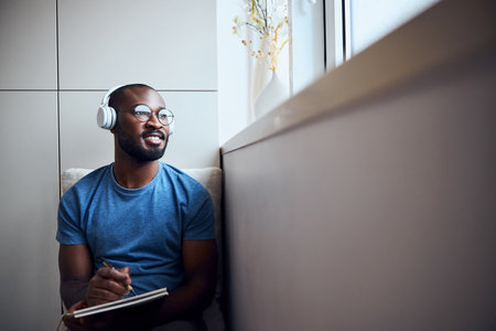 Multiracial artist in glasses lying on a couch and watching outside the window while using a pen