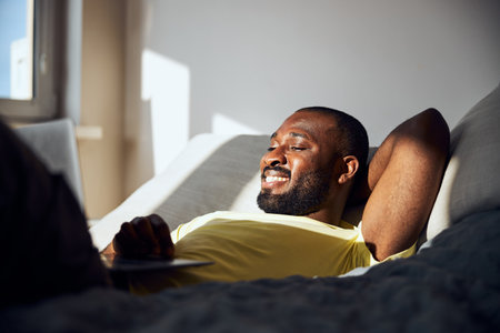 Male lying in bed with a sun shining on his face and having a laptop on his bellyの写真素材