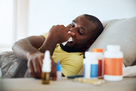Person stretching his arm from bed for a nasal spray while holding a tissue at his noseの写真素材