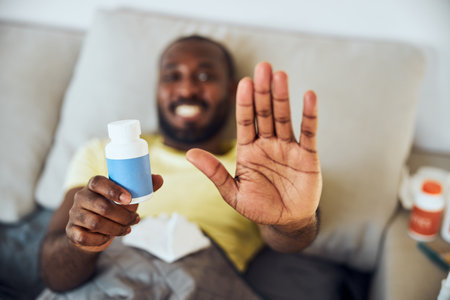 Cheerful sick adult showing a stop sign with palm of his hand while lifting a blue pack of medicineの写真素材