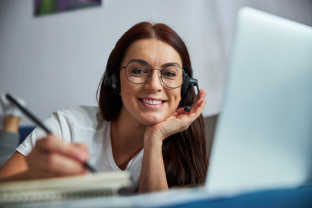 Smiling young woman in glasses is writing in sketchbook while enjoying communication in internet...