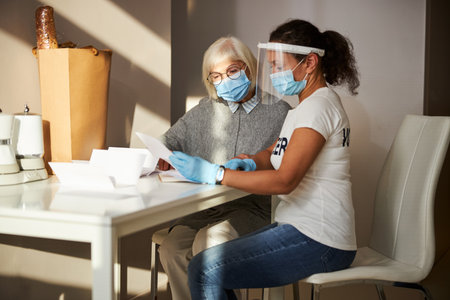 Senior lady in eyeglasses and a volunteer in a face shield perusing papers at the table
