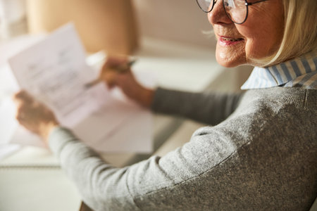 Cropped photo of a joyous blonde pensioner holding a sheet of paper and a penの写真素材