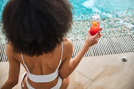 Copy-space photo of curly brunette woman in white bikini sitting by the swimming pool and holding a delicious drinkの写真素材